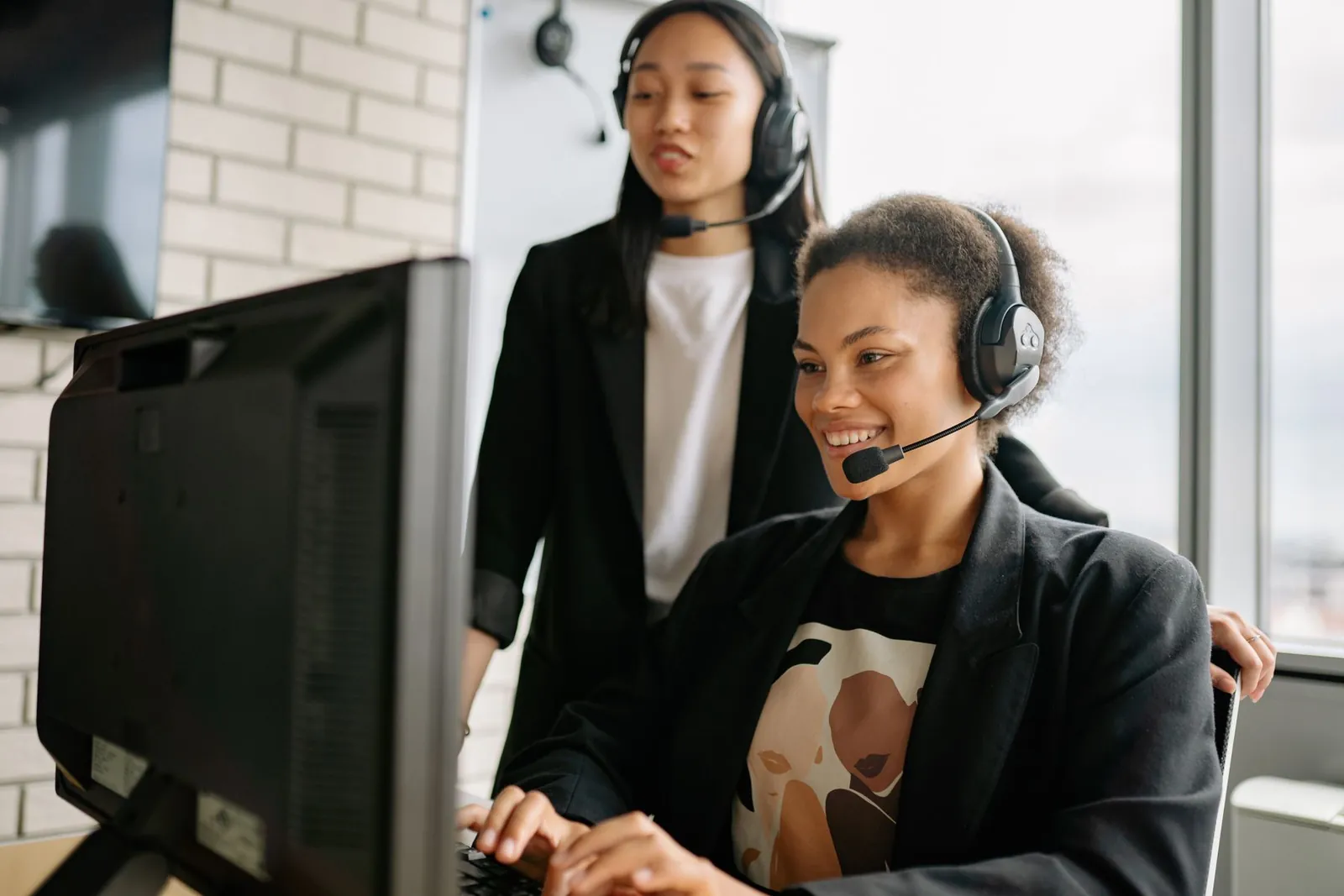 Contact centre supervisor standing beside a seated agent, coaching her while they look at the live wallboard on her screen